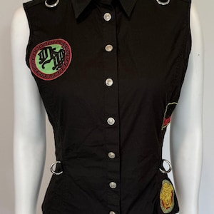 May include: Black sleeveless button-up shirt with silver buttons and metal ring accents. Features embroidered patches with logos in red, green, and gold. The shirt has a collared neckline and a fitted silhouette.