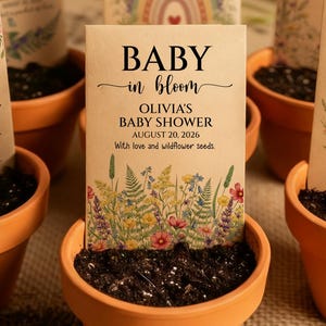 Puede incluir: Seis macetas de terracota llenas de tierra, cada una con un sobre de semillas. Los sobres presentan diseños florales y el texto "Baby in Bloom" con detalles de la fiesta de bienvenida del bebé. Algunos sobres tienen un arcoíris o una mariposa.