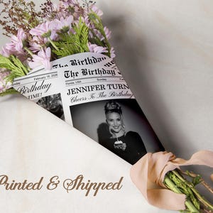 May include: A bouquet of pink flowers and greenery wrapped in a newspaper-style paper. The paper features the headline "The Birthday" and a black and white photo of a woman. A ribbon ties the bouquet.