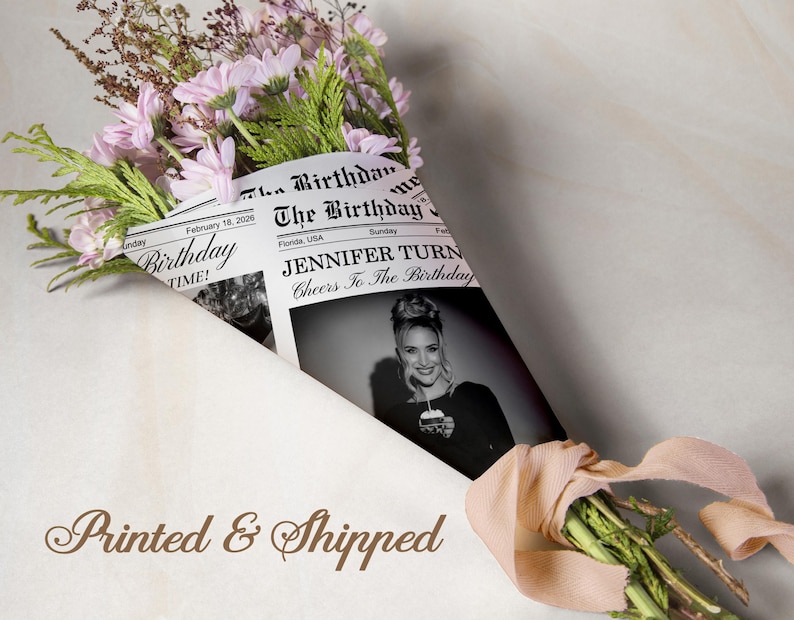 May include: A bouquet of pink flowers and greenery wrapped in a newspaper-style paper. The paper features the headline "The Birthday" and a black and white photo of a woman. A ribbon ties the bouquet.