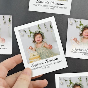 May include: A hand holding a white photo print with a baby girl wearing a floral crown and a light green dress. The text "Sophia's Baptism" and "THANKS FOR CELEBRATING WITH ME!" are printed on the photo, along with the date.