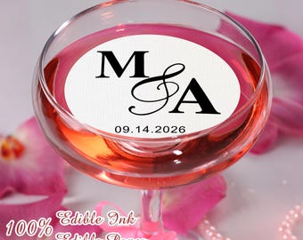 May include: A champagne glass filled with a pink liquid, featuring a white circular label with the letters "M & A" and the date "09.14.2026" in black. The glass is surrounded by pink flower petals and a pink ribbon.