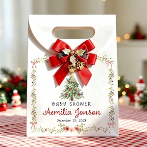 Op de afbeelding: Een witte papieren geschenktas met een rode satijnen strik, gouden belletjes en hulstblaadjes. De tas toont een kerstboom in aquarel, een bloemenrand en de tekst: Baby Shower, Aemilia Jonson, December 15, 2028, in het zwart en rood.
