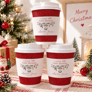 May include: Three red, ribbed disposable coffee cups with white lids and white sleeves. The sleeves feature the text 'a merry little BABY SHOWER', 'Aemilia Jonson', and '10/15/2028', along with a graphic of baby clothes hanging on a string.