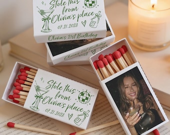 May include: White matchboxes with green text that reads "Stole this from Olivia's place 07.21.2028 Olivia's 21st Birthday." One box is open, revealing red-tipped matches and a photo. A lit candle and scattered matches are also visible.