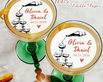May include: Two champagne glasses with green stems, each holding a cocktail with a circular edible paper topper. The toppers feature a champagne bottle pouring into a tower of glasses, with the names "Olivia & Daniel" and the date "04.21.2026".