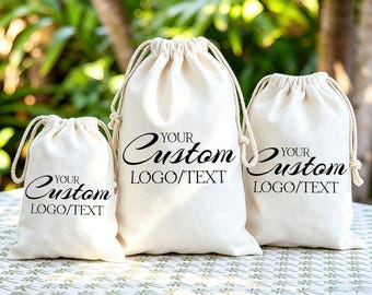 Könnte beinhalten: Drei cremefarbene Kordelzugbeutel in verschiedenen Größen, die jeweils mit dem schwarzen Text "YOUR Custom LOGO/TEXT" auf der Vorderseite bedruckt sind. Die Beutel bestehen aus hellem Stoff und haben passende Kordeln zum Verschließen.