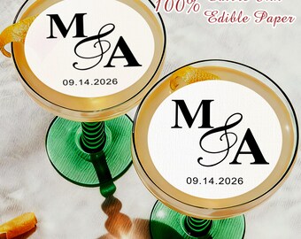 May include: Two coupe glasses with green stems, each filled with a light-colored drink and a citrus garnish. The drink's surface features a white edible paper circle with the letters "M & A" and the date "09.14.2026". Text reads "100% Edible Ink Edible Paper".