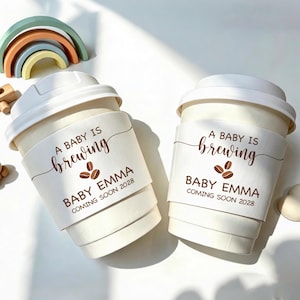May include: Two white paper coffee cups with white lids, each featuring a sleeve printed with the text A BABY IS brewing, BABY EMMA, and COMING SOON 2028, accompanied by a small coffee bean graphic. A pastel-coloured rainbow arch sits nearby.
