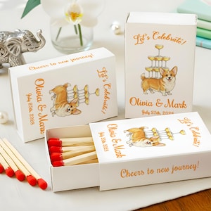 May include: White matchboxes with custom text and a corgi illustration. The boxes read "Let's Celebrate!" and "Cheers to new journey!" with the names "Olivia & Mark" and the date "July 25th, 2028".