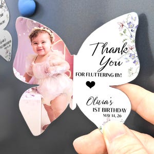 May include: Butterfly-shaped magnet with a photo of a baby girl in pink. Text on the magnet reads "Thank You" and "Olivia's 1st Birthday May 14, 26", with floral details. A magnet is held by a hand.