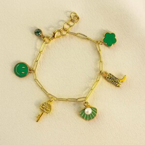 Puede incluir: Pulsera de dijes dorada con varios colgantes. Los dijes incluyen una carita sonriente verde, un trébol de cuatro hojas, una bota de vaquero, una llave, una concha con una perla y una pequeña gema verde. La pulsera está sobre una superficie color crema.