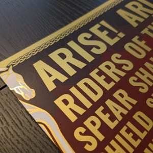 May include: A close-up of a decorative print with gold lettering on a burgundy background. The text reads "ARISE! RIDERS OF THEODEN..." with a horse head illustration. The print has a gold border with a braided design.