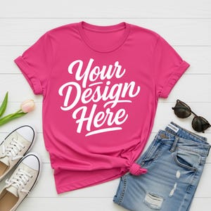 May include: A magenta t-shirt with the text "Your Design Here" in white script. The shirt is paired with light blue denim jeans, white sneakers, and black sunglasses. A pink tulip adds a touch of color.