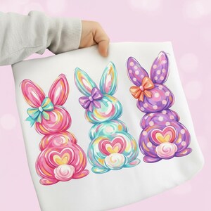 Może przedstawiać: Biała tkanina z trzema kolorowymi ilustracjami królików wielkanocnych. Każdy królik ma korpus w kształcie serca, kokardę i kropki. Króliki są różowe, niebieskie i fioletowe.