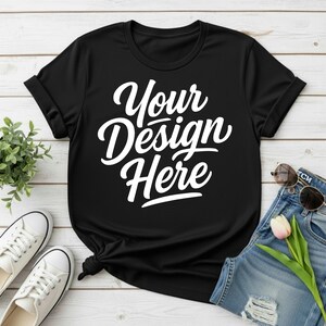 May include: Black t-shirt with the text "Your Design Here" in white script. The shirt is styled with rolled-up sleeves and a knotted hem. Accessories include white sneakers, sunglasses, and denim shorts with a flower.