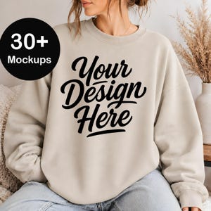 May include: A beige crewneck sweatshirt featuring the black text Your Design Here printed on the front. A black circular badge in the upper left corner displays the white text 30+ Mockups. The garment has long sleeves and a ribbed neckline.