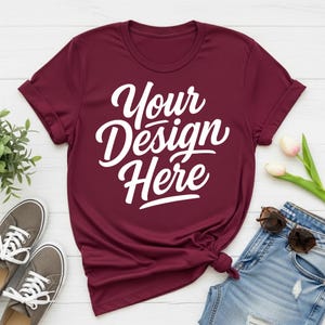 May include: A maroon t-shirt with the text "Your Design Here" in white script. The shirt is styled with rolled-up sleeves and a knot at the bottom. Gray sneakers and blue jeans with sunglasses are also in the image.