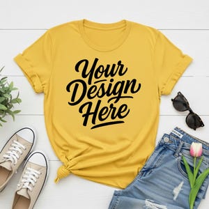 May include: Mustard yellow t-shirt with the text "Your Design Here" in black script. The shirt is styled with blue jeans, sunglasses, and sneakers, suggesting a casual, everyday look.