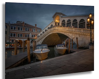 May include: A photograph of the Rialto Bridge in Venice, Italy, at dusk. Two small boats are docked in the canal. The bridge is lit up with lights, and there are people on the bridge. The sky is a dark blue.