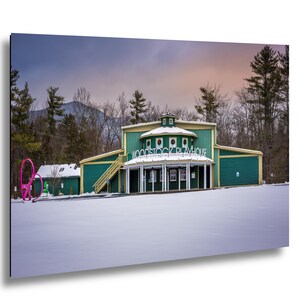 Puede incluir: Una escena invernal del Woodstock Playhouse, un edificio verde con adornos amarillos. Las palabras "Woodstock Playhouse" están sobre la entrada. Una escultura rosa se encuentra en primer plano, con árboles y una montaña al fondo.