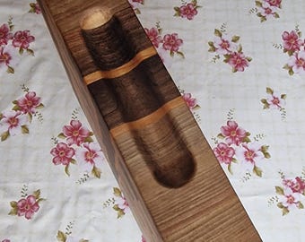 Puede incluir: Un soporte de madera hecho a mano para botellas de vino. El soporte es rectangular con un interior tallado para sostener una botella y dos copas. La madera presenta tonos marrones claros y oscuros, con una banda oscura. Mide unos 30 cm.
