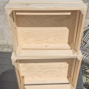 May include: Two rectangular light-colored wooden crates stacked vertically. Each crate features a slatted design with visible natural wood grain, reinforced corner supports, and open sides, constructed from unfinished light pine wood planks.