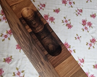 Puede incluir: Un soporte de madera para botellas de vino con un interior tallado para sostener una botella y un soporte para tapones incorporado. El soporte está hecho de varios tonos de madera y se encuentra sobre una superficie con estampado floral.