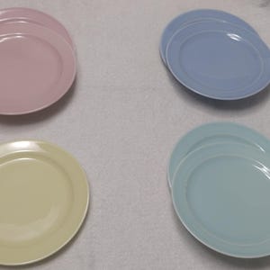 May include: Four sets of ceramic plates in pastel colors: pink, light blue, yellow, and teal. Each set includes two plates. The plates have a simple, round design with a slightly raised rim, suitable for serving food.