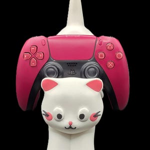 Op de afbeelding: Een witte controllerstandaard in de vorm van een kat die een roze en zwarte draadloze gamecontroller vasthoudt. Het katfiguur heeft roze oren, roze wangmarkeringen en zwarte ogen, neus en mond. De controller is voorzien van standaardknoppen en joysticks.