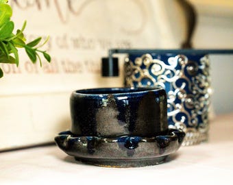 Puede incluir: Un cenicero de cerámica azul oscuro con una base a juego. El cenicero tiene un acabado brillante y una apariencia única, hecha a mano. Un recipiente decorativo azul y plateado está en el fondo.