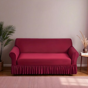 May include: A claret-coloured sofa cover with a frilled skirt. The cover is fitted over a sofa with curved arms. A potted plant and a small side table with decorative items are in the background. The room has a neutral colour palette.