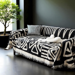 Könnte beinhalten: Eine schwarz-weiß gemusterte Sofadecke mit geometrischem Design. Der Überwurf ist über ein Sofa drapiert, mit einem passenden Kissen und dekorativen Gegenständen. Die Decke hat einen Fransenbesatz und ist aus einem weichen Material gefertigt.