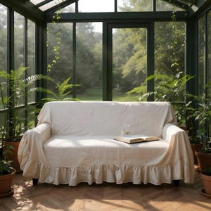 May include: A cream-colored sofa with a ruffled skirt is placed in a sunroom filled with potted plants. An open book and a glass of water sit on the sofa. The room has large windows and a view of the outdoors.