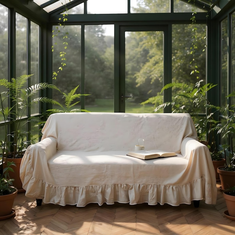 May include: A cream-colored sofa with a ruffled skirt is placed in a sunroom filled with potted plants. An open book and a glass of water sit on the sofa. The room has large windows and a view of the outdoors.