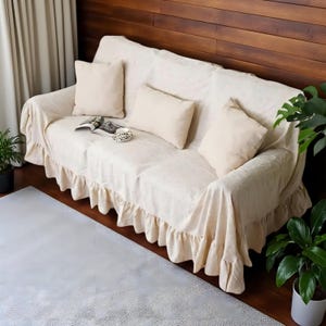 May include: A cream-colored sofa is draped with a matching cover and three throw pillows. The cover has a ruffled skirt detail. A book and a decorative cup sit on the sofa. Potted plants are on either side of the sofa.