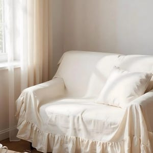 May include: Cream-colored sofa with a matching throw and pillow. The sofa is covered with a ruffled slipcover, creating a soft, inviting aesthetic. The room is bathed in natural light, enhancing the cozy atmosphere.
