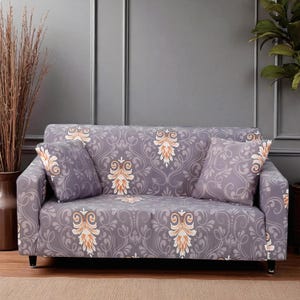 以下が含まれることがあります： 白とオレンジの花柄のグレーのソファ。お揃いのクッションが2つソファにあります。ソファは黒い脚で、灰色の壁に面しています。