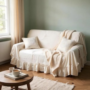 May include: A cream-colored sofa with a ruffled skirt, covered with a matching slipcover and two throw pillows. A cream-colored knitted throw blanket is draped over the arm. A small wooden table holds a cup of coffee and a book.