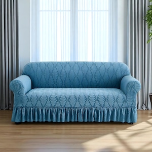May include: A light blue sofa cover with a leaf pattern and a ruffled skirt. The cover is draped over a sofa, with the arms and back covered. The background includes a window with sheer curtains and a wooden floor.