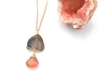 May include: A gold chain necklace with a green and black teardrop-shaped pendant and a smaller pink teardrop-shaped pendant. The necklace is on a white surface next to a pink crystal.