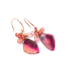 May include: A pair of dangle earrings featuring watermelon tourmaline gemstones. The earrings have a rose gold wire and hook design. The gemstones are a mix of pink and red hues, with smaller pink beads clustered above the larger stones.