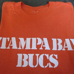 May include: Orange t-shirt with white text that reads "Tampa Bay Bucs".