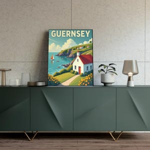 May include: A framed art print featuring a coastal scene with the word "GUERNSEY" at the top. The artwork depicts a white house with a red roof, a sailboat, and a green hillside. The print is displayed on a dark green cabinet with gold legs.