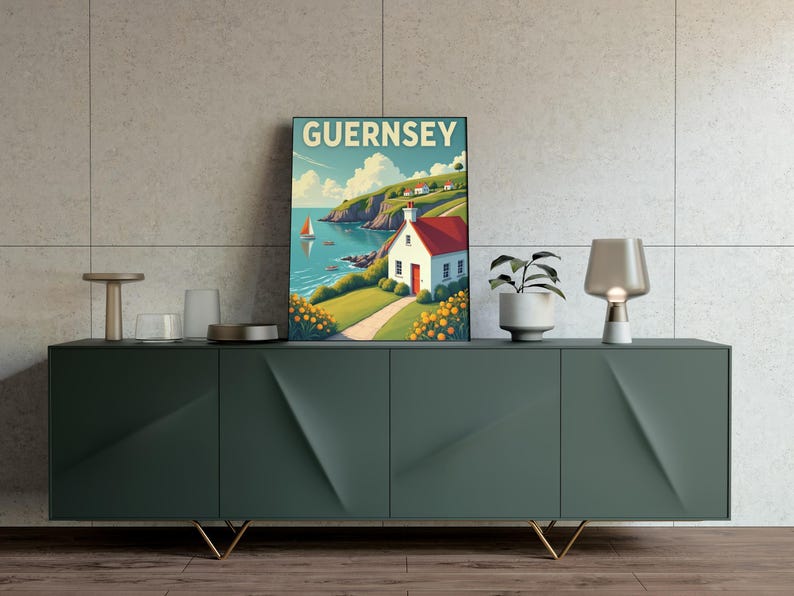 May include: A framed art print featuring a coastal scene with the word "GUERNSEY" at the top. The artwork depicts a white house with a red roof, a sailboat, and a green hillside. The print is displayed on a dark green cabinet with gold legs.