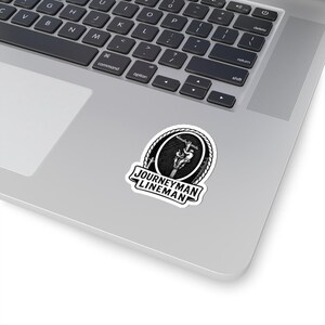 May include: A black and white sticker with the text "JOURNEYMAN LINEMAN". The sticker shows a lineman working on a power line inside a circular frame with a rope border. The sticker is on a laptop. The laptop is silver.
