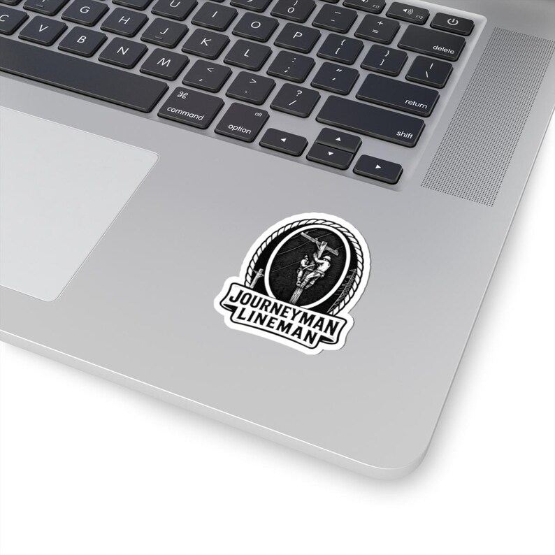 May include: A black and white sticker with the text "JOURNEYMAN LINEMAN". The sticker shows a lineman working on a power line inside a circular frame with a rope border. The sticker is on a laptop. The laptop is silver.