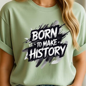 Puede incluir: Camiseta verde salvia con el texto "BORN TO MAKE HISTORY" en una fuente audaz y estilizada. El texto está sobre un diseño abstracto en negro y gris. La camiseta tiene mangas enrolladas.