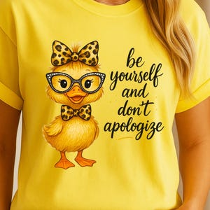 May include: Yellow t-shirt featuring a cartoon duck wearing glasses and a bow tie with a leopard print pattern. The text "be yourself and don't apologize" is written in black script on the shirt.
