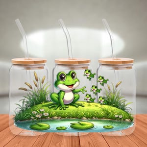 Peut inclure: Trois gobelets en verre avec couvercles en bambou et pailles transparentes. Chaque gobelet présente un dessin de grenouille de dessin animé avec une scène d'étang. Les grenouilles sont vertes avec des ventres blancs, et la scène comprend des nénuphars et de l'herbe.